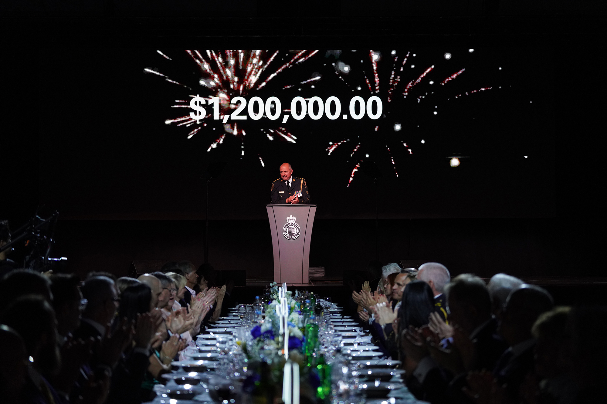 Chief Jim MacSween addresses a crowd at PAN, the screen behind him shows $1.2 million dollars was raised for charity